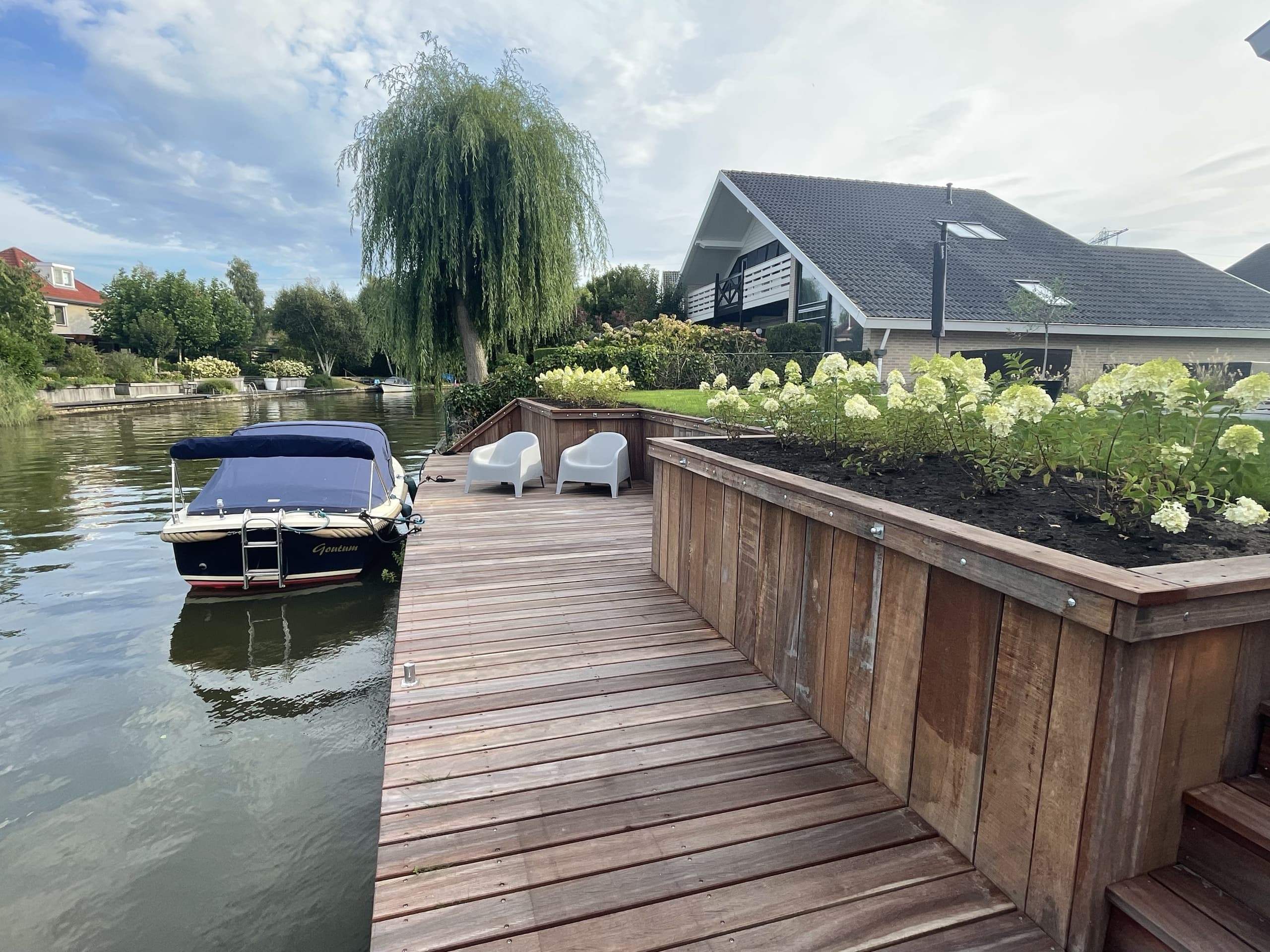 De afgewerkte vlonder met plantenbakken vol hortensia's, twee stoelen en de aangemeerde boot, met de vaart ervoor