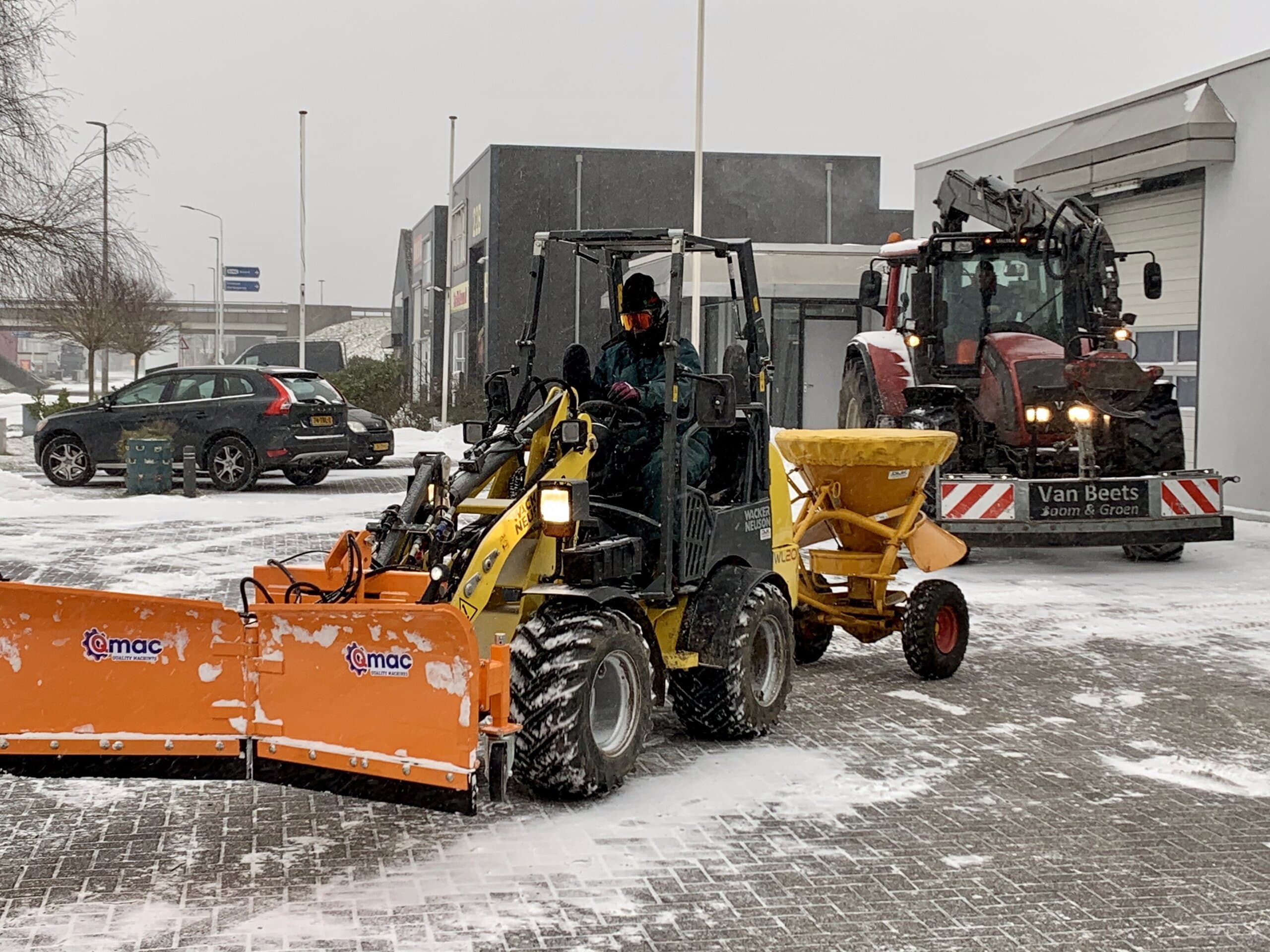 Shovel met oranje sneeuwschuif en gele zoutstrooier ruimt een besneeuwd bedrijventerrein, met daarachter een tractor van Van Beets met strooiwagen
