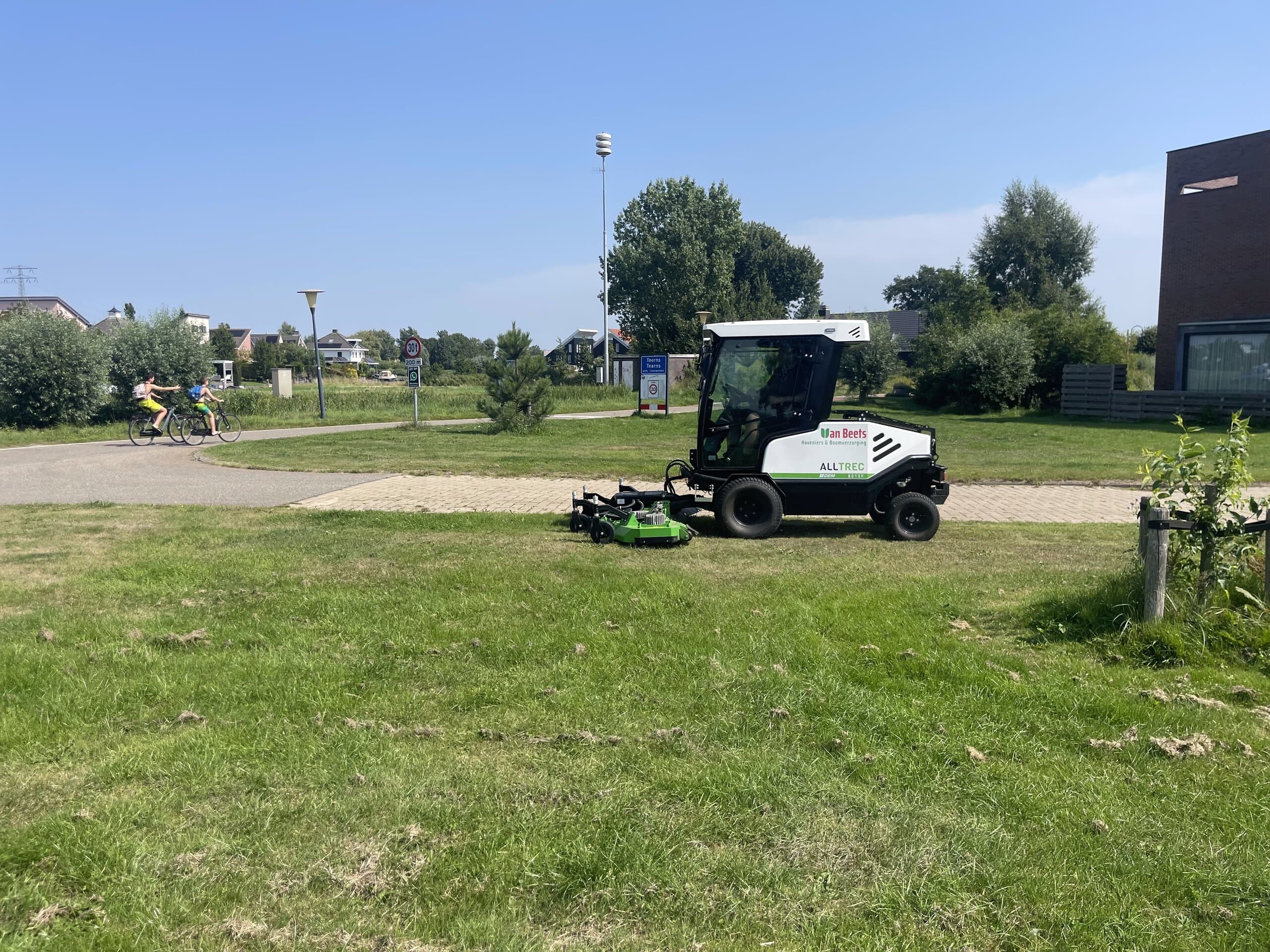 Elektrische maaimachine van Van Beets maait een grasveld in de openbare ruimte, met fietsers op het pad erachter