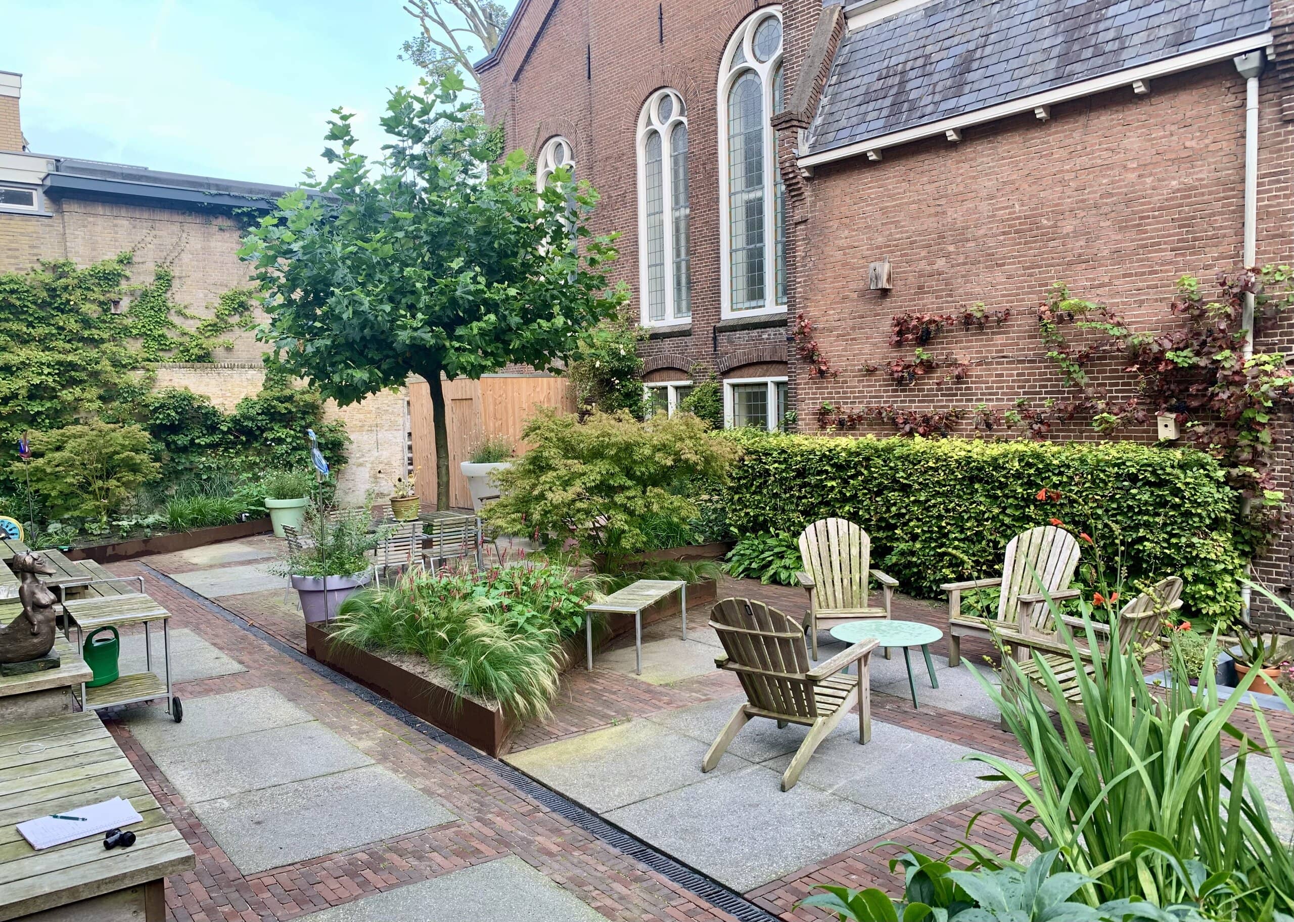 Stadstuin in de binnenstad van Leeuwarden met een plataan, houten stoelen, beukenhaag en borders met siergrassen langs een oude kerkgevel